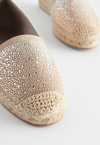 Béžové espadrilky s perforovaným textilným zvrškom, ktorý obsahuje metalické akcenty. Jutový a pletený povrch okolo podrážky dodáva textúru.