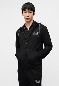 Μαύρο μπουφάν με φερμουάρ και κουκούλα, κατασκευασμένο από μαλακό ύφασμα. Διαθέτει λογότυπο στο στήθος, μπροστινές τσέπες και ριμπ μανσέτες. Συνδυάζεται με παντελόνια σετ.