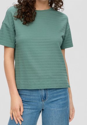Kvinde iført en grøn, ribbet t-shirt med korte ærmer og blå jeans, med krøllet hår delvist synligt mod en enkelt grå baggrund.