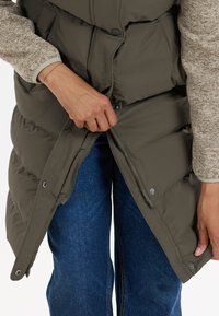 Olivgrüne Steppweste mit Druckknopf- und Reißverschluss, getragen über einem hellbraunen strukturierten Pullover. Unten sind blaue Jeans sichtbar.
