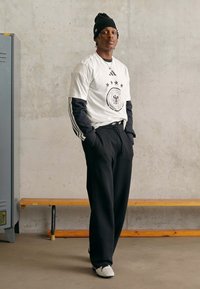 Mann i svart lue, hvite lagdelte treningskjorter, svarte joggebukser og hvite joggesko, står med hendene i lommene ved siden av en trebenn.