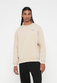 Beżowy polarowy sweatshirt z okrągłym dekoltem, ściągaczami przy rękawach i na dole, z małą kolorową naszywką na piersi. Czarne spodnie.