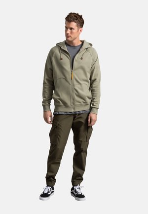 Felpa con zip verde chiaro dotata di cappuccio, progettata con cuciture piatte. Abbinata a pantaloni cargo verde oliva e sneakers in bianco e nero.
