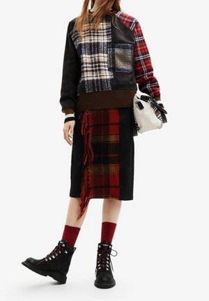 Personne portant un pull en patchwork à carreaux et une jupe assortie à carreaux avec des franges, des chaussettes rouges, des bottes noires à lacets, tenant un sac à main blanc.