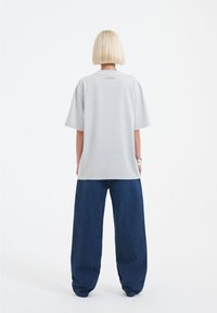 Šedé oversized tričko s krátkými rukávy, hladkou texturou a malým růžovým logem na zadní straně, v kombinaci s širokými tmavě modrými džíny.