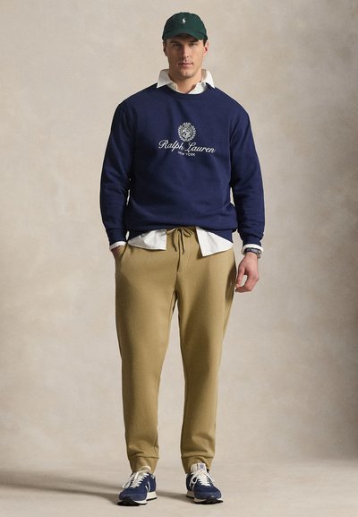 Man draagt een marineblauwe Ralph Lauren sweater, een wit overhemd met kraag, beige joggers, een marineblauwe pet en blauwe sneakers, staand tegen een neutrale achtergrond.
