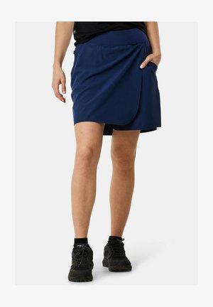 Vrouw draagt een marineblauwe wikkelrok met split, zwarte sneakers en zwarte enkelsokken, staat met één hand in de zak.