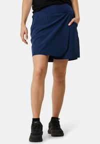 Navy blue skort made from lightweight fabric, featuring a wrap design, a side pocket, and paired with black athletic shoes.