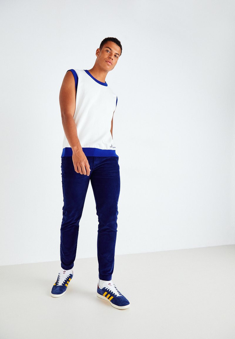 Top blanc sans manches avec des accents bleus, associé à un pantalon en velours côtelé bleu marine et des baskets bleu marine avec des rayures jaunes. Design simple, texture lisse.