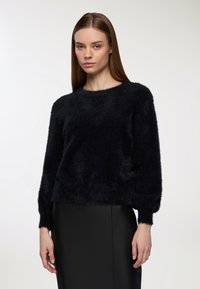 Czarny puszysty sweter z okrągłym dekoltem i długimi rękawami. Materiał wygląda na miękki i teksturowany, zestawiony z gładką czarną spódnicą.