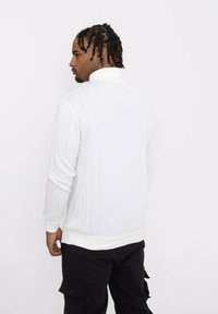 Pull en tricot blanc avec un col haut, présentant un motif texturé et des poignets et ourlet blancs contrastants, associé à un pantalon cargo noir.