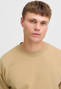 Beige särk ümmarguse kaelusega ja ribiga varrukatega, valmistatud pehmest kangast, sileda tekstuuri ja minimalistliku disainiga.