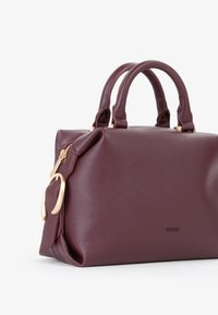 Borsa in pelle bordeaux con superficie liscia, doppi manici rotondi e dettagli in metallo dorato. Presenta una chiusura con zip e un logo impresso.