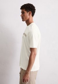 Helles cremfarbendes T-Shirt mit kurzen Ärmeln aus weichem Material, das ein bedrucktes Design auf der Vorderseite aufweist. Der Schnitt ist lässig mit einem runden Halsausschnitt.