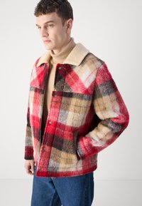 Plaid wool jacket in red, beige, and black with a shearling collar. Features snap buttons and side pockets, showcasing a textured fabric.