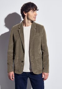 Blazer de pana de color verde oliva con dos bolsillos frontales, cierre de un solo botón y solapas muesca, usado sobre una camiseta de color beige claro y pantalones vaqueros azules.