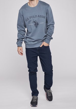 Mężczyzna ubrany w niebieską bluzę U.S. Polo Assn., ciemne dżinsy i szare trampki, stojący z jedną ręką w kieszeni na jednolitym tle.