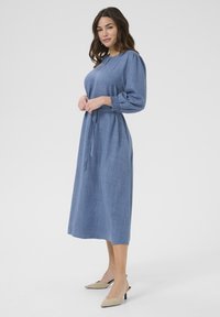 Robe bleue à manches longues en tissu texturé. Présente une taille cintrée avec une ceinture assortie et une longueur jusqu'au genou. Chaussures de couleur neutre.