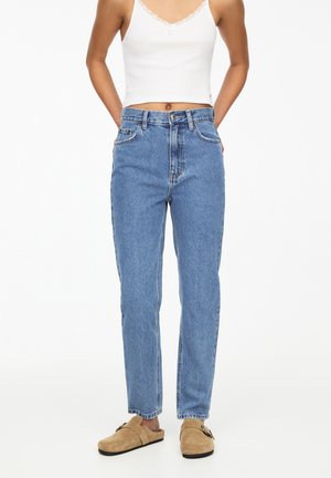 Femme portant un jean bleu taille haute, un haut blanc sans manches et des chaussures beiges à enfiler, debout les mains dans le dos.