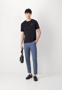 Svart kortärmad t-shirt med liten vit logotyp, i kombination med ljusblå skräddarsydda byxor, svart väska och svarta loafers.