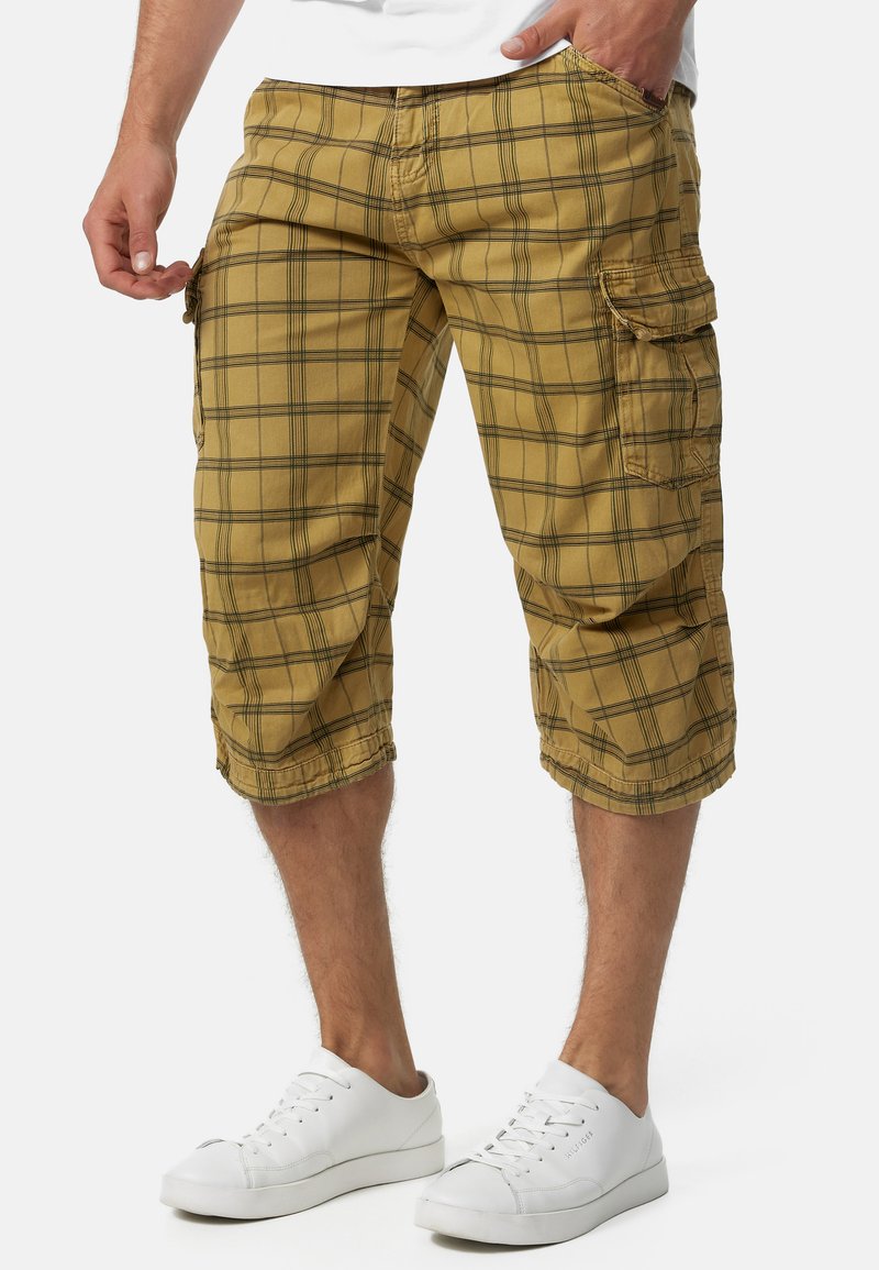 Shorts cargo à carreaux bruns avec des lignes noires, dotés de poches latérales et d'une coupe décontractée. Portés avec des baskets blanches sur un fond clair.