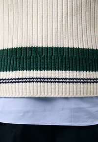 Sweter dziany w kremowym kolorze z ciemnozielonymi i granatowymi paskami na dole, z teksturowanym wzorem prążkowanym. Nałożony na jasnoniebieską koszulę.