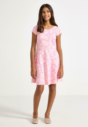 Hymyilevä tyttö seisoo päällään lyhythihainen, vaaleanpunainen kukkamekko ja vaaleat pitsiset balettitossut yksiväristä valkoista taustaa vasten.