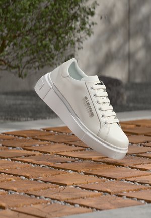 Valentino valged paeladega tossud, külgedel metallist logo, hõljuvad mustrilise puidupinna kohal, taustal rohelus.