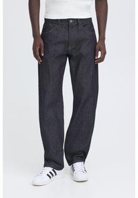 Mörkblå jeans med raka ben, använda av en stående person med vita sneakers och en vit topp, händerna avslappnade vid sidorna.