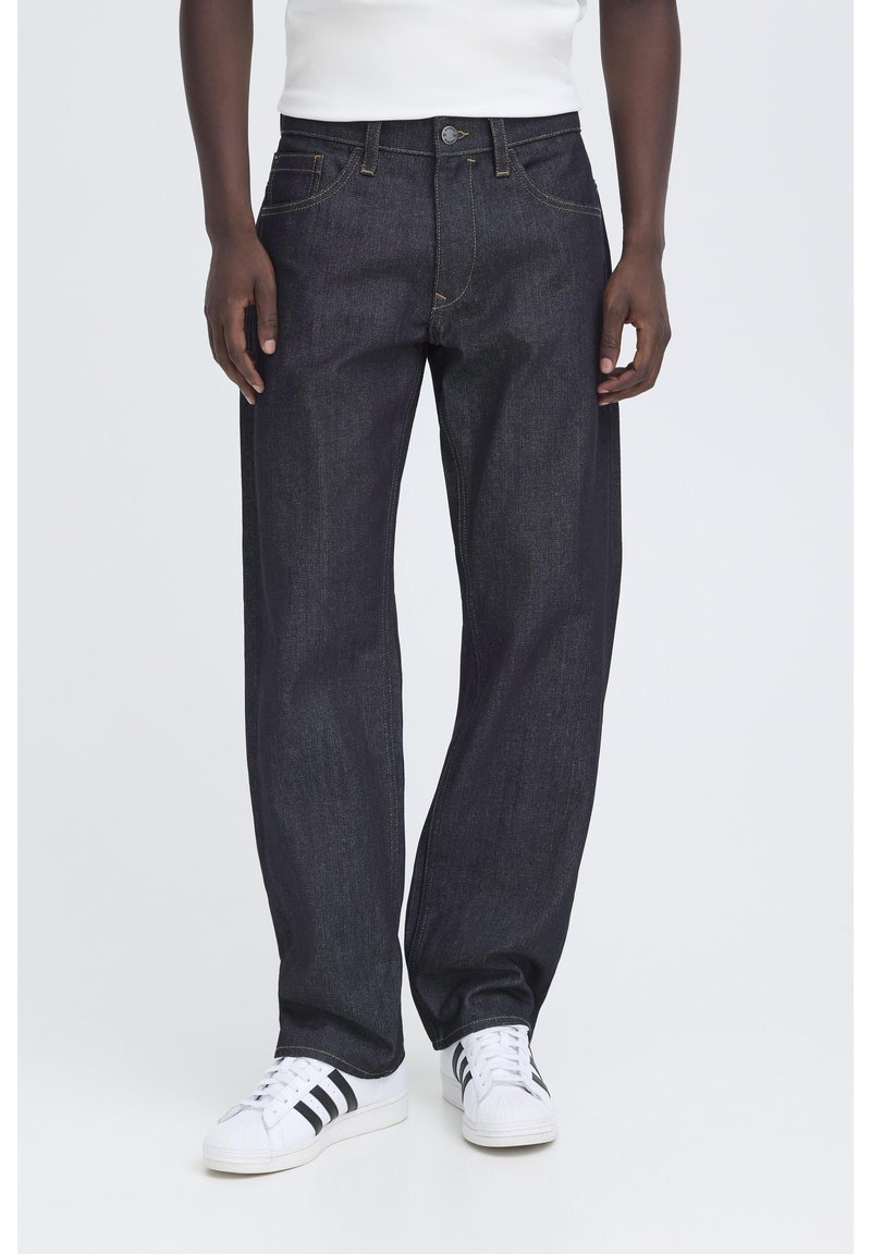 Mörkblå jeans med raka ben, använda av en stående person med vita sneakers och en vit topp, händerna avslappnade vid sidorna.