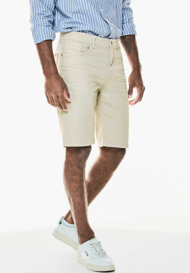 Béžové kraťasy s hladkou texturou, spárované s světle modrou pruhovanou košilí a bílými teniskami. Kraťasy mají poutka na opasek a přední kapsy.