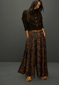 Hnědý krátký svetr se zapínáním na knoflíky, kombinovaný s dlouhou leopardí sukní s plisovaným vzorem a kapsami; nošený s béžovými teniskami.