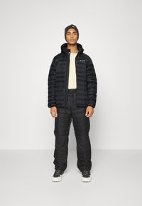 Giacca a vento nera con cappuccio e chiusura a zip, abbinata a pantaloni isolanti coordinati. Sotto indossa una maglietta chiara.