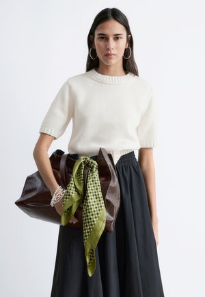 Frau mit glattem dunklem Haar, die einen weißen kurzärmligen Pullover und einen schwarzen Rock trägt, hält eine große braune Ledertasche mit einem grünen gemusterten Schal.