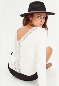 Pull blanc avec un dos en V, présentant des détails de dentelle. Le mannequin porte un chapeau noir et est assis devant un fond neutre.