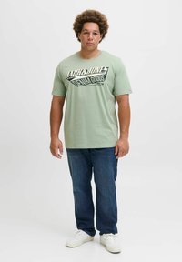 Tricou scurt din bumbac, de culoare verde deschis, cu text grafic negru; asortat cu blugi de culoare albastru închis și adidași albi. Design simplu și casual.