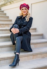 Abrigo de lana negro con botones, pantalones de cuero negro ajustados y botas de tacón alto brillantes negras. Boina roja con un patrón texturizado.