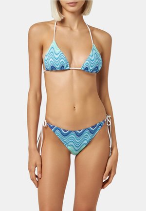 Donna con un bikini a stringhe dal motivo ondulato blu e verde con lacci bianchi, in piedi davanti a uno sfondo bianco uniforme.