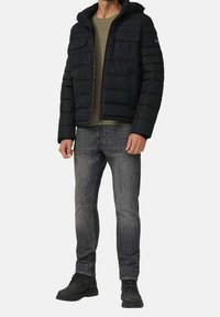 Zwarte pufferjack met een capuchon en een rits aan de voorkant, met twee borstzakken, gedragen over een groene gebreide trui en grijze jeans. Zwarte laarzen.