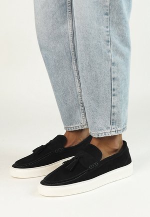 Zwarte suede tassel loafers met witte rubberen zolen, gedragen met lichtblauwe cropped denim jeans op blote voeten.