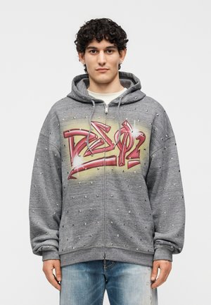 Mladý muž kučeravých vlasov nosí sivú mikinu na zips s cvokmi, s červeným graffiti nápisom na hrudi a svetlomodrými džínsami.