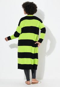 Cardigan lungo a strisce nere e verde neon con polsini e orlo a costine. Indossato sopra leggings neri e sandali leggeri. Texture in maglia morbida.