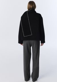 Abrigo negro de gran tamaño con un destacado detalle de costura gris, combinado con pantalones de pierna ancha grises. El abrigo cae de forma asimétrica sobre el hombro.