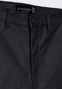 Jean en denim noir avec une texture lisse, dotés d'une fermeture à bouton, de passants de ceinture et d'une étiquette affichant la taille et les informations sur la marque.