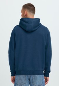 Homme avec les cheveux courts et une boucle d'oreille portant un sweat à capuche bleu marine et un jean bleu clair, de dos face à un fond clair.