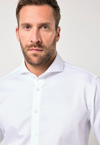 Bílá košile s jemným texturovaným vzorem, s klasickým límečkem a knoflíkovou légou, vystavená na neutrálním pozadí.