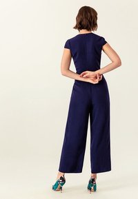 Námořnicky modrý overal s krátkými rukávy, širokými nohavicemi a zapínáním na zip. Kombinován s barevnými podpatky s geometrickým vzorem.