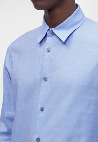 Chemise bleu clair à boutons avec un petit motif répétitif de texte blanc et un col classique portée par une personne.