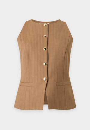 Femme portant un haut marron sans manches boutonné, un jean bleu à motifs, des chaussures beiges et un sac à bandoulière marron à franges.