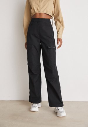 Osoba ubrana w czarne spodnie typu cargo z logo Calvin Klein Jeans, beżową skróconą kurtkę oraz białe sneakersy, stojąca na beżowej podłodze.